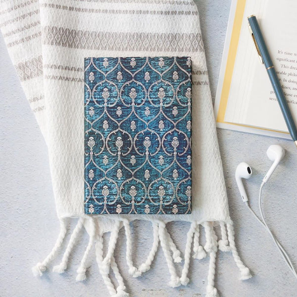 En blå bok med silverfärgat mönster på pärmen. Den ligger på en filt tillsammans med penna och hörlurar. Boken finns i serien Blue Velvet från Paperblanks - Tidformera