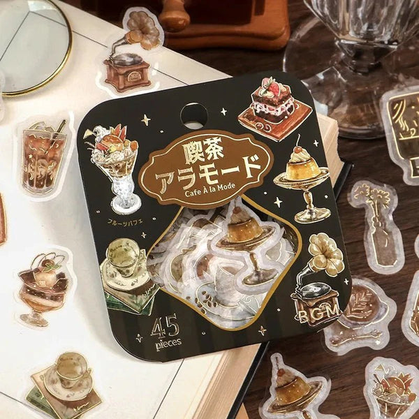 Serier - Coffee a la mode från BGM - En förpackning klistermärken ligger på en anteckningsbok. Runt skrivboken ligger små flake stickers utströdda. Motiven är med drinkar och bakelser. Tidformera