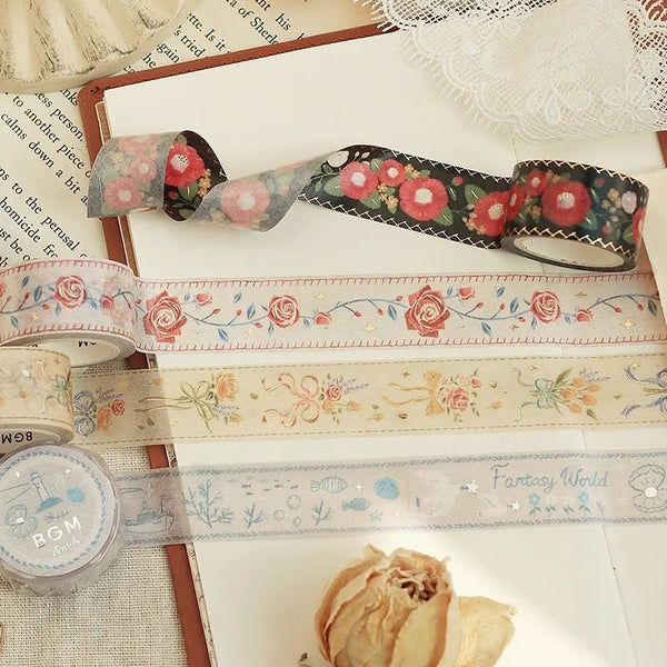Serier - Embroidered Ribbon från BGM. Fyra rullar washi tape ligger utrullade på en anteckningsbok. Motiven på dekorationstejpen är blommor som är broderade. Tidformera