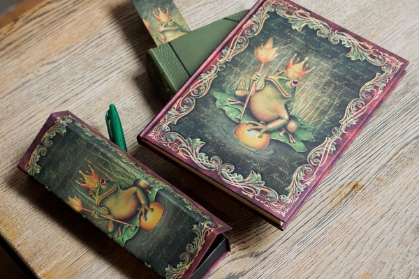 Pennskrin, anteckningsbok och bokmärke ur serien Fairy tale collection från Paperblanks. Motivet är en groda med trollstav och krona på huvudet från Bröderna Grimms Grodprinsen - Tidformera
