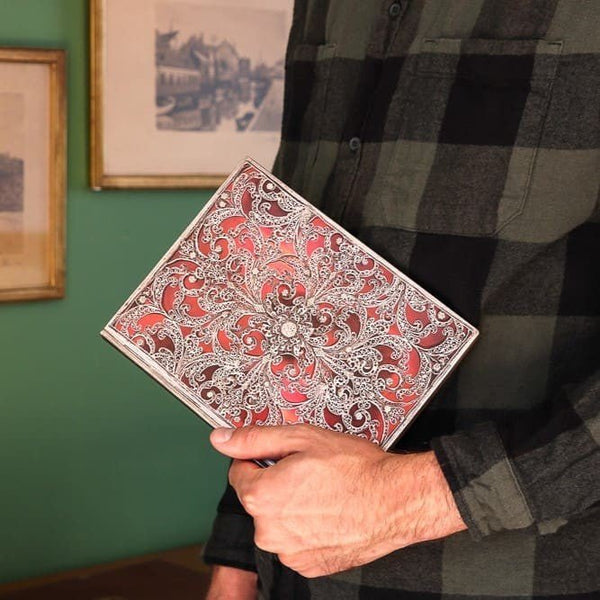 En man som håller i en skrivbok i serien Garnet från Paperblanks. En röd bok med silver mönster. - Tidformera