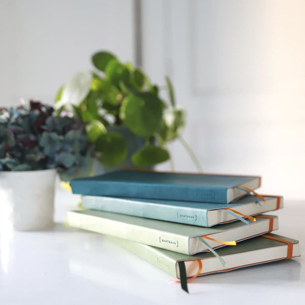 Serier - Goalbook från Rhodia. En hög med fyra gröna och blåa böcker ligger på ett bord. I bakgrunden är det krukor med blommor.  - Tidformera