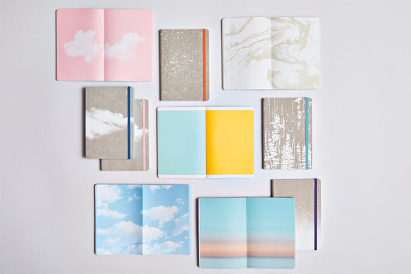Anteckningsböcker ur serien Inspiration Book från Nuuna - Flera skrivböcker ligger utspridda och uppslagna. Motiv i är med moln, strand, soluppgång och enfärgat i gult och blått. Tidformera