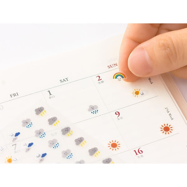 Serier - Kalenderstickers trackers från Midori - Stickersark ligger på en kalender. På stickersarket är det olika moln och sol. En hand sätter in en regnbåge i kalendern. Tidformera