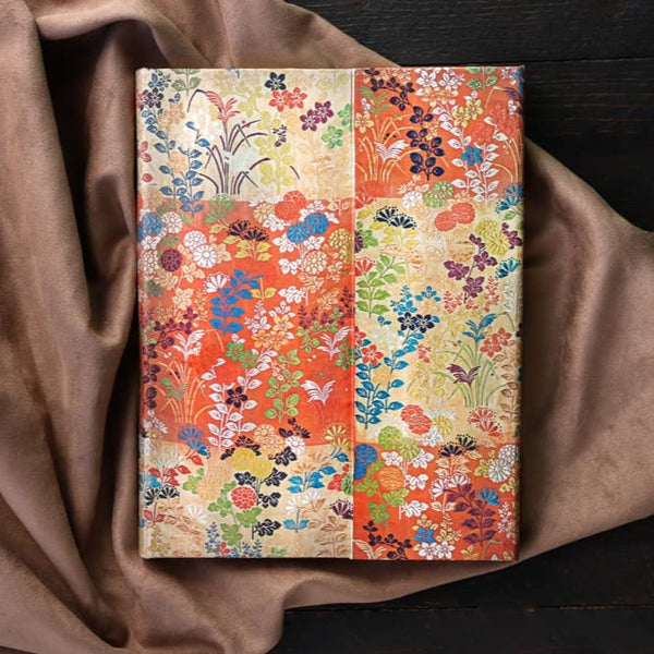 Anteckningsbok i serien Kara-ori från Paperblanks. Skrivbok med blomstermönster ligger på en beigefärgad filt. Tidformera