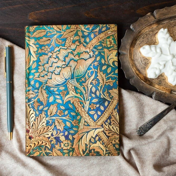 Anteckningsbok med motivet Morris Windrush ligger på ett bord. Skrivboken är turkos med guldpräglat mönster med stor blomma. Bredvid bok ligger en penna och en skål. Tidformera