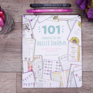 101 Layouter till din Bullet Journal från Tukan. Bok med den titeln ligger på ett bord. Bredvid är pennor och blommor - Tidformera