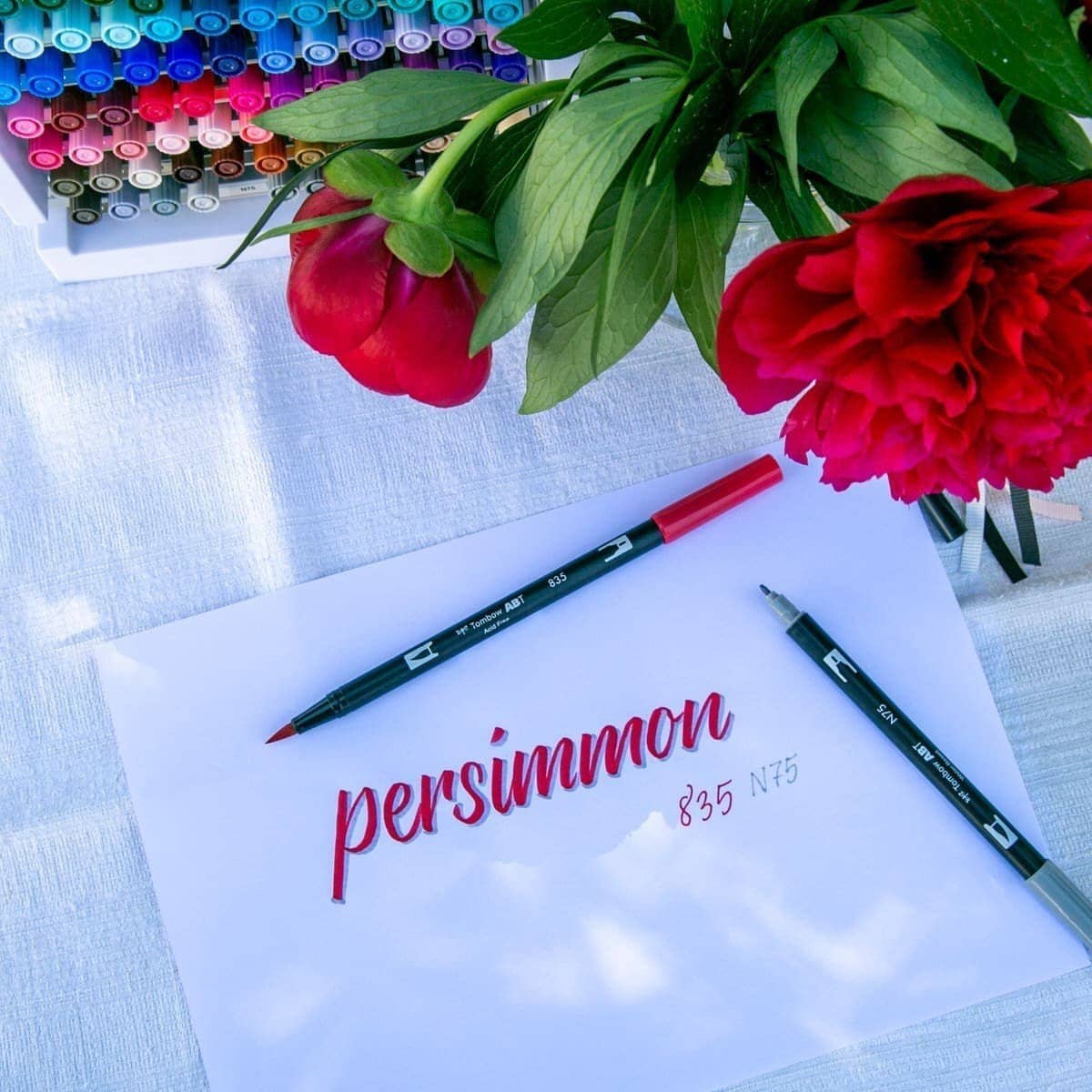 ABT Dual Brush pen - 835 Persimmon från Tombow. Röd och grå penna som använts att skriva namnet på den röda pennan. Tombows pennförvaring och blommor står också på bordet. - Tidformera