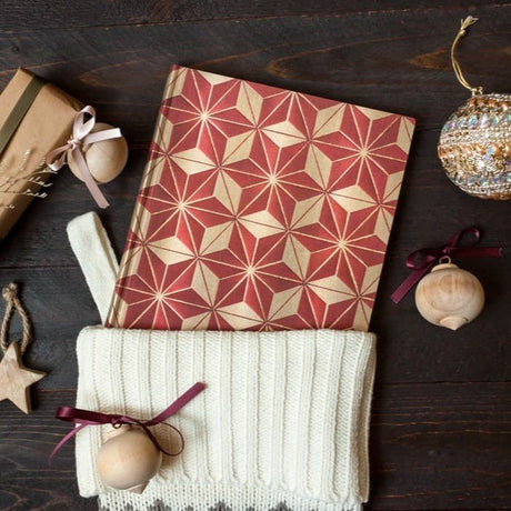 Röd och guldmönstrad anteckningsbok Hishi Midi Linjerad Flexis från Paperblanks i en julstrumpa. Bredvid finns julgranskulor. - Tidformera