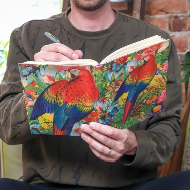 En man skriver i sin färggranna bok som har papegoja och blommor på båda fram- och bakpärm. Anteckningsbok Tropical garden Ultra Linjerad från Paperblanks - Tidformera