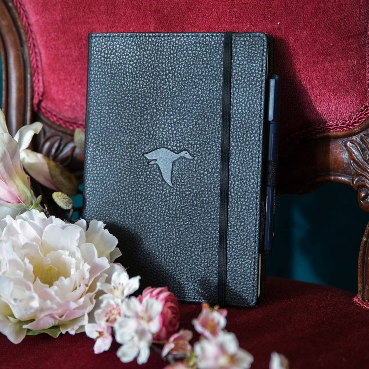Svart anteckningsbok Wildlife Dotted A5 Black Duck från Dingbats* . Boken står på en stol med röd sammet. Framför  boken finns det skira blommor.- Tidformera