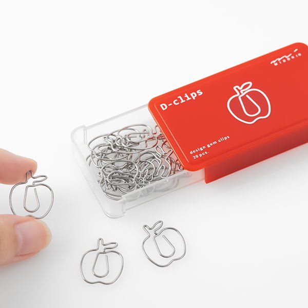 D - clips Gem - Apple från Midori. Eb förpackning med gem är öppnad och fingrar håller i gem som är formad som ett äpple.  Tidformera