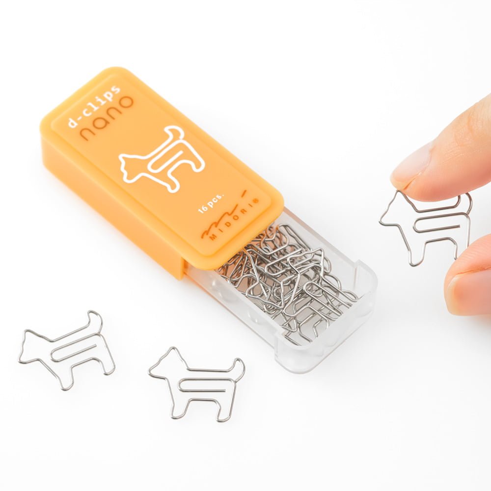 D - clips Nano Gem - Dog från Midori. En öppnad gul förpackning med små gem. Fingrar håller i ett superlitet gem som är formad som en hund.  Tidformera