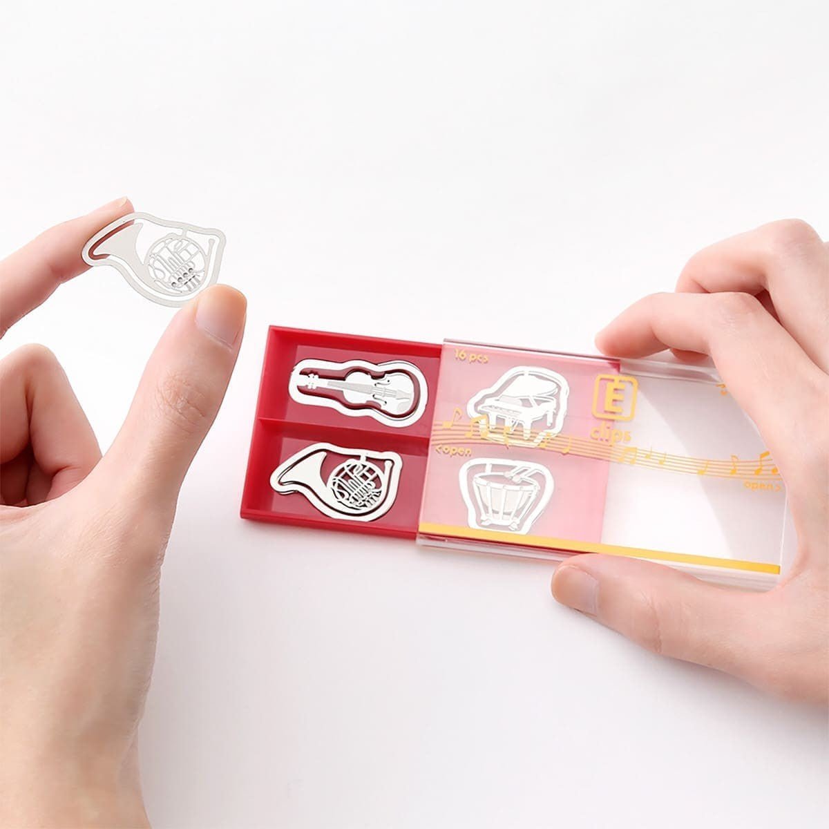 E - clips Gem i metall - Musical instruments från Midori. En hand håller i förpackningen med gem och lyft ut ett gem med motiv med ett blåsinstrument. Tidformera