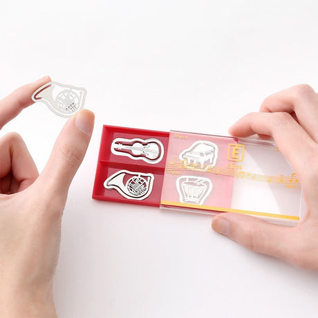 E - clips Gem i metall - Musical instruments från Midori. En hand håller i förpackningen med gem och lyft ut ett gem med motiv med ett blåsinstrument. Tidformera