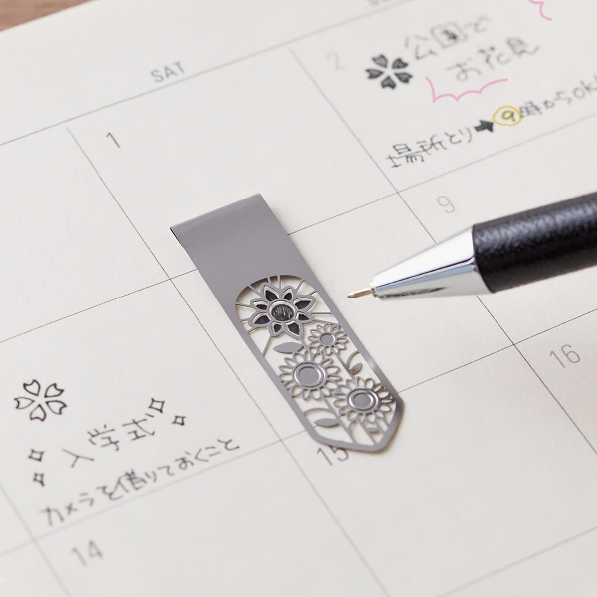 Ett bokmärke ligger på en kalender. En penna ritar i stencilen på bokmärket. - Clip Bookmarker Flower från Midori.