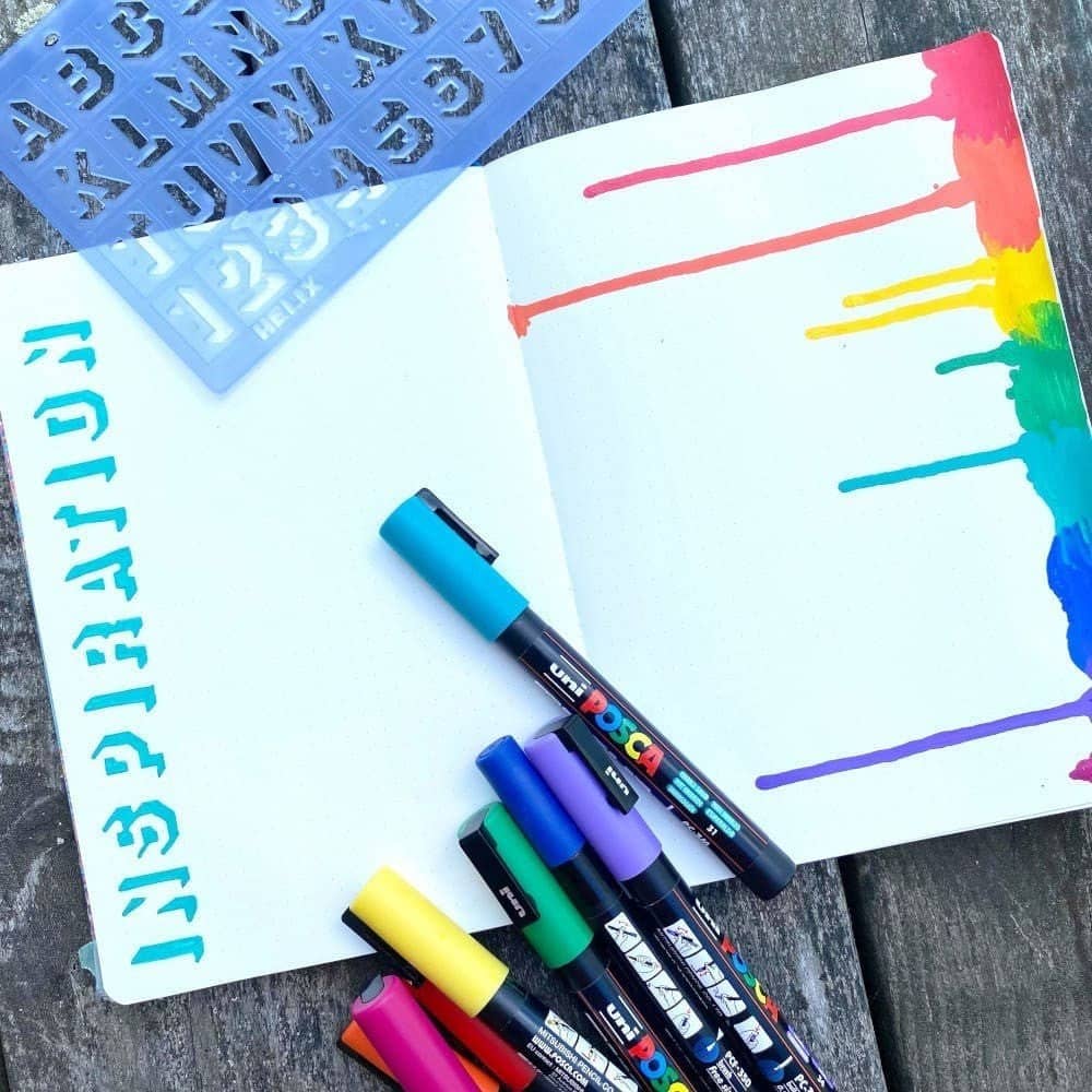 En uppslagen anteckningsbok med pennor i flera färger. Stencil ligger på skrivbok och använts till att skriva Inspiration.