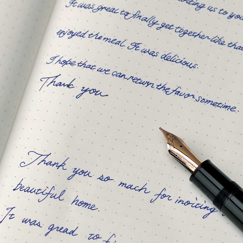 TomoeRiver Notebook softcover - A5 - Dot Grid från Sakae TP. En uppslagen anteckningsbok med dotted inlaga. Reservoarpenna ligger på  skrivboken och har skrivit med blått bläck. i skrivboken. Tidformera