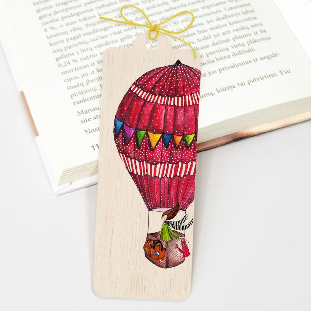 Wooden bookmark - Fly Away från Cozywood. Ett rektangulärt bokmärke med en luftballong ligger på en läsebok. Bokmärket har ett gult snöre. Tidformera