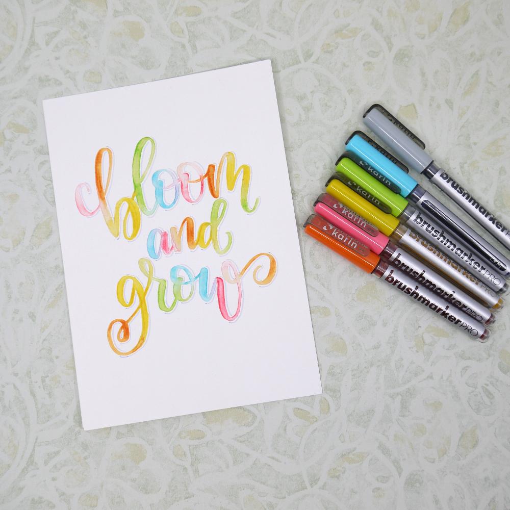 Brushmarker Pro pennor ligger bredvid ett papper med texten Bloom and grow. Texten är skrivet med brush lettering och de pennorna.