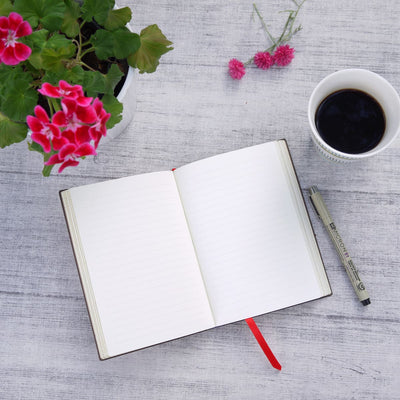 En linjerade skrivbok ligger uppslaget på ett bord. Det finns även en penna, kaffekopp och blommor på bordet.- Tidformera