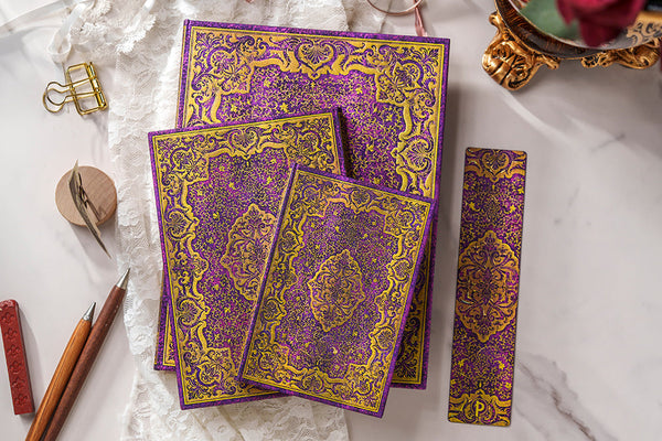 Hög med anteckningsböcker i olika format och bokmärke. Serien Picaresque från Paperblanks. Motiv är mönster i lila och guld. - Tidformera