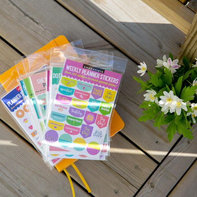 Serier - Planner stickers Peter Pauper Press - Flera förpackningar med stickers ligger i en hög på en gul anteckningsbok. Bredvid högen är det en vas med vitsippor. Tidformera