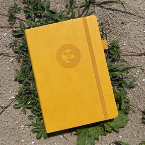 Dotted Notebook A5 160 GSM Smiling sun från Paper24. Gul anteckningsbok med motiv med en leende sol. Skrivboken ligger ute i solen på växter. - Tidformera